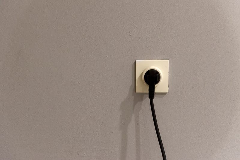 A black plug inserted into a white wall socket on a plain grey wall